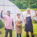 収穫した野菜を持つ笑顔の女性を中央に両隣に笑顔の男性が立っています